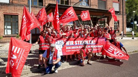 Rund zwanzig Personen stehen vor einem Backsteingebäude und halten rote Fahnen sowie ein großes rotes Banner. Auf den Fahnen und Warnwesten sind EVG-Logos sichtbar. Das Banner zeigt den Schriftzug „WIR STREIKEN!“ sowie weiteren kleineren Text. Die Personen tragen überwiegend rote Warnwesten und stehen dicht beieinander am Straßenrand bei sonnigem Wetter.