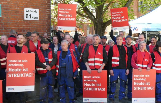 Mehrere Personen stehen vor einer Backsteinwand neben einem Schild mit der Aufschrift „Gebäude 61 Nord“. Viele tragen rote Warnwesten und blaue Arbeitskleidung. Mehrere Personen halten rote Schilder mit der weißen Aufschrift „Heute ist kein Arbeitstag. Heute ist STREIKTAG!“ und „EVG“-Logo. Im Hintergrund sind ein Baum, ein blaues Zelt und weitere Personen zu sehen.
