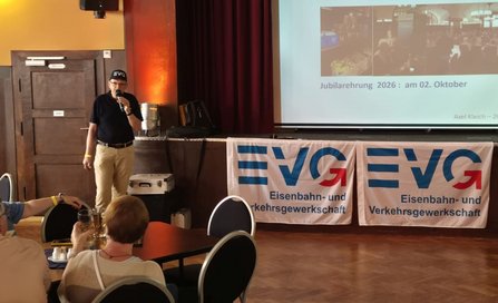 Innenraum mit Bühne und Projektionsfläche. Eine Person mit Mikrofon steht vor zwei Bannern mit der Aufschrift „EVG Eisenbahn- und Verkehrsgewerkschaft“. Vor der Bühne sitzen Personen an Tischen. Auf der Projektionsfläche ist unter anderem der Text „Jubilarehrung 2026: am 02. Oktober“ zu erkennen.