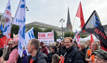 Menschen stehen dicht gedrängt auf einem Platz mit vielen Fahnen und Transparenten. Mehrere weiß-blaue Fahnen mit dem Schriftzug „Verkehrs-“ sind sichtbar. Im Hintergrund steht ein Kirchturm mit Uhr sowie weitere Gebäude. Ein Transparent trägt den Text „EURE KRIEGE NICHT AUF UNSEREN RÜCKEN!“.