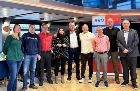 Gruppenfoto in einem Innenraum mit Holzfußboden und großen Fensterflächen. Neun Personen stehen nebeneinander in einer Reihe vor zwei Roll-up-Bannern mit dem sichtbaren Schriftzug „EVG“. Die Personen tragen überwiegend Freizeit- und Business-Kleidung in verschiedenen Farben, darunter Grün, Blau, Rot, Schwarz, Rosa und Grau. Im Hintergrund sind Tische und Stühle zu sehen. Die Aufnahme ist frontal und zeigt die Personen von Kopf bis Fuß.