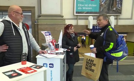Ein Informationsstand in einer Bahnhofshalle mit mehreren Personen davor. Auf dem Tisch liegen Flyer, Klemmbretter und Becher. Eine Person steht hinter dem Tisch, zwei weitere Personen stehen davor; mehrere Gesichter sind verpixelt. Eine Person trägt eine Tasche mit dem gut lesbaren Text „I AM MADE FOR MORE“. Im Hintergrund sind Bahnhofswände, Durchgänge und Plakate sichtbar.