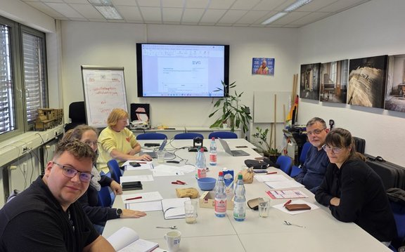 Besprechungsraum mit einem rechteckigen Tisch, an dem sechs Personen sitzen. Auf dem Tisch liegen Unterlagen, Notizbücher, Stifte, Mobiltelefone, Gläser, Tassen, Wasserflaschen, eine Schüssel und kleine Papiertüten. An der Wand befindet sich ein großer Bildschirm mit einer geöffneten Dokumentansicht, daneben ein Flipchart mit handschriftlichen Notizen. Rechts stehen Zimmerpflanzen, mehrere Stäbe sowie Wandbilder. Links sind Fenster mit Jalousien und ein Modell eines Waggons auf der Fensterbank zu sehen. Die Personen arbeiten mit Laptops und Papierunterlagen.