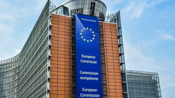 Modernes Bürogebäude mit geschwungener Glasfassade. An der Front hängt ein großer blauer Banner mit gelben Sternen im Kreis und den Texten „#TeamJunckerEU“, „Europese Commissie“, „Commission européenne“ und „European Commission“. Darüber ist ein runder Gebäudeteil mit Glas- und Metallelementen zu sehen, im Hintergrund ein heller Himmel mit Wolken.