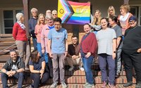 Gruppenfoto auf einer Treppe vor einem Gebäude mit Veranda und Türen im Hintergrund. Etwa 16 Personen stehen, sitzen oder hocken auf den Stufen. In der oberen Mitte wird eine mehrfarbige Regenbogenfahne mit der Aufschrift „EVG *queer“ gehalten. Die Fahne zeigt zusätzlich farbige Streifen in Pfeilform auf der linken Seite. Mehrere Personen tragen Freizeitkleidung in verschiedenen Farben. Die Szene ist bei hellem Sonnenlicht im Freien aufgenommen.