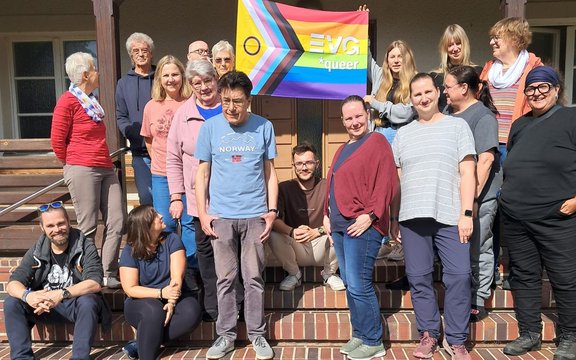 Gruppenfoto auf einer Treppe vor einem Gebäude mit Veranda und Türen im Hintergrund. Etwa 16 Personen stehen, sitzen oder hocken auf den Stufen. In der oberen Mitte wird eine mehrfarbige Regenbogenfahne mit der Aufschrift „EVG *queer“ gehalten. Die Fahne zeigt zusätzlich farbige Streifen in Pfeilform auf der linken Seite. Mehrere Personen tragen Freizeitkleidung in verschiedenen Farben. Die Szene ist bei hellem Sonnenlicht im Freien aufgenommen.