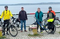 Vier Personen stehen nebeneinander auf einem gepflasterten Weg vor einem Metallgeländer an einem See. Jede Person trägt einen Fahrradhelm und steht neben oder hält ein Fahrrad; mehrere Fahrräder mit Gepäckträgern und Taschen sind sichtbar. In der Mitte des Vordergrunds steht ein kurzer Betonpoller. Hinter dem Geländer ist eine ruhige Wasserfläche zu sehen, dahinter eine bewaldete Uferlinie. 