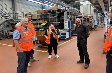 Mehrere Personen in orangefarbenen Warnwesten stehen in einer großen Werkhalle. Im Hintergrund sind Regale mit Kabeln, Kartons, Werkzeugwagen, Maschinen und eine Metalltreppe zu sehen. Der Boden ist mit gelb markierten Laufwegen versehen.