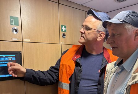 Zwei Personen mit dunklen Kappen stehen vor einem in Holzwand eingelassenen Touchscreen. Eine Person zeigt auf den Bildschirm. Beide tragen orangefarbene Warnwesten. Über dem Bildschirm sind Schrankfächer mit Piktogrammen, darunter ein Erste-Hilfe-Symbol.