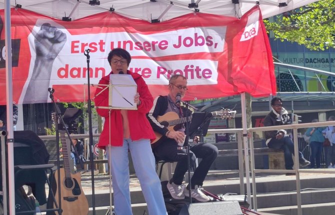 Kleine überdachte Bühne im Freien mit zwei Personen an Mikrofonen. Eine Person steht mit einem Blatt oder Ordner vor einem Notenständer, eine weitere Person sitzt mit einer akustischen Gitarre. Hinter der Bühne hängt ein rotes Banner mit der Aufschrift „Erst unsere Jobs, dann eure Profite“ sowie „DGB“. Im Hintergrund sind Bäume, Stufen und ein Gebäude zu sehen.