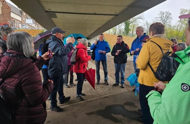 Unter einer Straßenbrücke steht eine Gruppe von Personen in einem Halbkreis zusammen. Die Personen tragen wetterfeste Jacken und halten teilweise Regenschirme oder Taschen. Die Gesichter sind unscharf dargestellt. Im Hintergrund sind Brückenpfeiler, ein gepflasterter Boden und angrenzende Bebauung zu sehen.