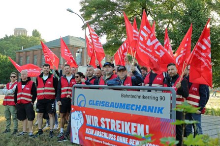 Gruppe von Personen in roten Warnwesten versammelt sich im Freien hinter einem Schild mit der Aufschrift „Bahntechnikerring 90“ und „GLEISBAUMECHANIK“. Mehrere rote Fahnen mit dem Schriftzug „EVG“ werden über der Gruppe gehalten. Vor dem Schild befindet sich ein rotes Banner mit dem Text „WIR STREIKEN! KEIN ABSCHLUSS OHNE GEWERKSCHAFT“. Im Hintergrund stehen Bäume, ein Backsteingebäude und eine Straßenleuchte.