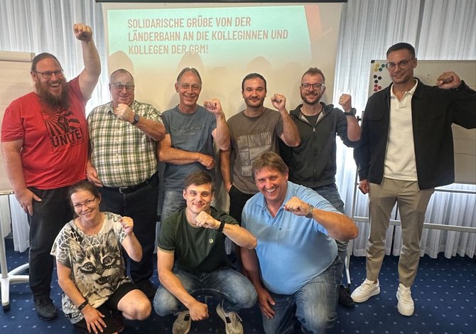 Gruppenfoto in einem Innenraum vor einer Leinwand mit projiziertem Text. Neun Personen stehen oder knien in zwei Reihen und halten jeweils einen angewinkelten Arm mit geballter Faust nach oben. Im Hintergrund sind Vorhänge, ein Flipchart und ein Whiteboard sichtbar. Auf der Leinwand steht: „SOLIDARISCHE GRÜßE VON DER LÄNDERBAHN AN DIE KOLLEGINNEN UND KOLLEGEN DER GBM!“.