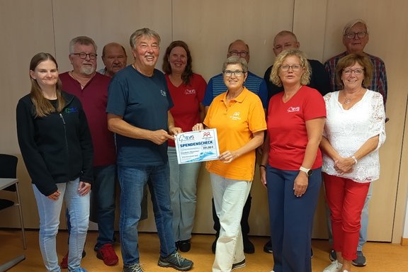 Elf Personen stehen für ein Gruppenfoto in einem Raum. In der Mitte halten zwei Personen gemeinsam einen Spendenscheck in die Kamera. Die Gruppe ist vor einer hellen Schrankwand aufgestellt. Fünf Personen stehen in der hinteren Reihe, sechs Personen in der vorderen Reihe. Die Kleidung ist überwiegend sportlich und Freizeitkleidung in verschiedenen Farben, darunter Dunkelblau, Rot, Orange, Weiß und Schwarz.