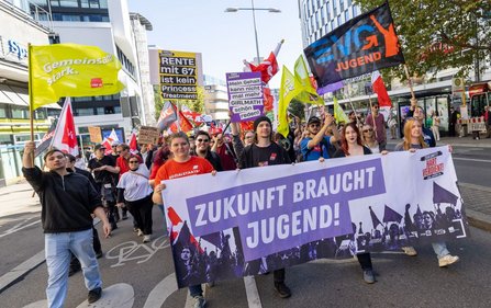 Demonstrationszug auf einer breiten Straße. Mehrere Personen tragen Fahnen und ein großes rotes Banner mit der Aufschrift „Hart verdient! Deine Arbeit. Deine Gesundheit. Deine Rente.“. Im Hintergrund sind Bäume, Gebäude und weitere Fahnen zu sehen.