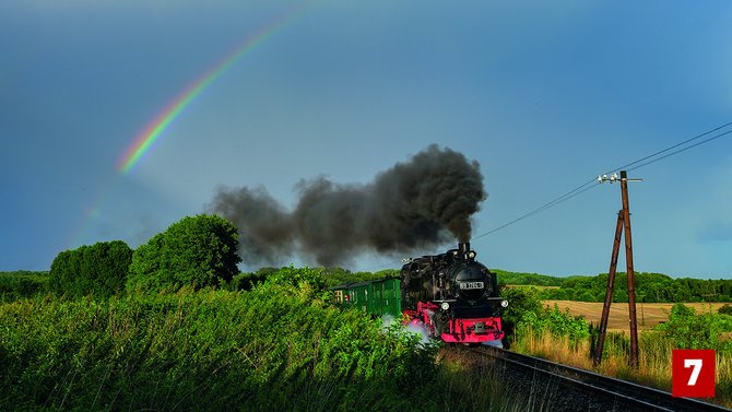 Schwarze Dampflokomotive mit grünen Personenwagen auf einer eingleisigen Bahnstrecke in einer ländlichen Landschaft. Dichte schwarze Rauchwolken steigen aus dem Schornstein auf. Links im Himmel ist ein Regenbogen vor dunklen Wolken zu sehen. Die Strecke verläuft zwischen hohem Gras, Feldern und Bäumen. Rechts stehen Holzmasten mit Leitungen. Unten rechts befindet sich eine rote Markierung mit der Zahl „7“.