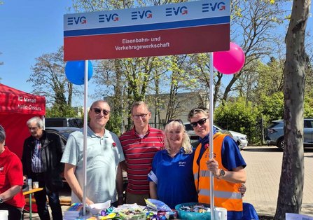 Vier Personen stehen hinter einem Infostand mit einem rechteckigen Schild, auf dem mehrfach „EVG“ und „Eisenbahn‑ und Verkehrsgewerkschaft“ steht. Am Stand liegen Broschüren, Süßigkeiten und kleine Gegenstände. Links und rechts sind bunte Luftballons befestigt. Im Hintergrund stehen weitere Personen, ein rotes Zelt mit der Aufschrift „IG Metall Halle‑Dessau“ sowie parkende Autos und Bäume.
