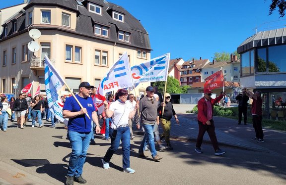 Straßenumzug mit mehreren Personen, die Fahnen tragen, auf einer asphaltierten Straße zwischen Wohnhäusern. Sichtbar sind Fahnen mit den Aufschriften „EVG“, „Eisenbahn- und Verkehrsgewerkschaft“ und „ver.di“. Die Personen gehen in einer Gruppe von links nach rechts; im Hintergrund stehen mehrstöckige Gebäude, Gehwege und weitere Teilnehmende. Blauer Himmel und Tageslicht.
