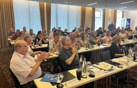 Seminar- oder Tagungsraum mit mehreren Reihen von Tischen. Zahlreiche Personen sitzen an den Tischen und klatschen. Auf den Tischen stehen Wasserflaschen, Gläser, Becher, Unterlagen und Namensschilder. Große Fenster mit Vorhängen befinden sich entlang der linken Raumseite.