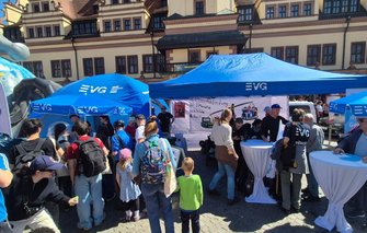 elebter Platz mit mehreren blauen Pavillons und Sonnenschirmen mit der Aufschrift „EVG“. Zahlreiche Menschen, darunter auch Kinder, stehen davor oder gehen vorbei. Auf einem Pavillon hängt ein Banner mit Text und Zeichnungen. Im Hintergrund steht ein historisches Gebäude.