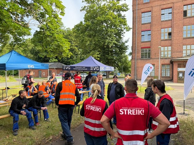 Mehrere Personen treffen sich draußen vor einem Gebäude zu einer Streikaktion, einige tragen Westen mit der Aufschrift „STREIK!“ und sitzen oder stehen unter EVG‑Zelten. Die Szene zeigt einen Außenbereich mit Bäumen und einem großen Backsteingebäude im Hintergrund. Mehrere Zelte mit der Aufschrift „EVG – Eisenbahn- und Verkehrsgewerkschaft“ sind aufgebaut. Darunter sitzen und stehen Gruppen von Personen an Bierzeltbänken. Einige tragen orange Warnwesten, andere rote Westen mit der deutlich sichtbaren Aufschrift „STREIK!“. Auf den Tischen liegen Gegenstände wie Taschen oder Getränke. In der Mitte unterhalten sich mehrere Personen im Stehen, während andere auf den Bänken sitzen. Rechts ist zusätzlich ein weißes Fahnenbanner mit EVG-Logo aufgestellt. Der Boden besteht aus Gras und einem schmalen Weg.