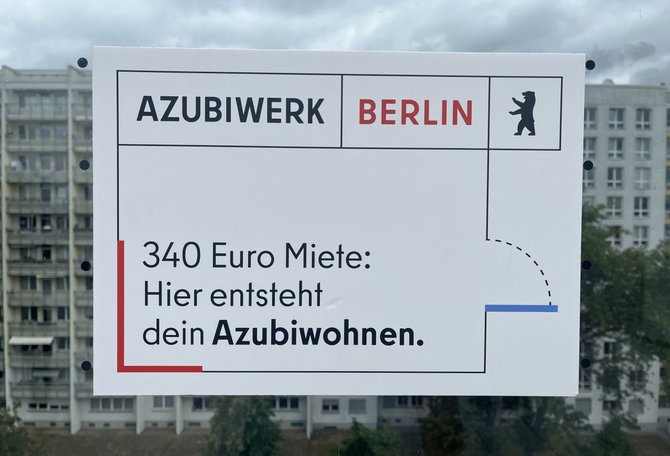 Weißes Schild vor mehrstöckigen Wohngebäuden und bewölktem Himmel. Auf dem Schild steht: „AZUBIWERK BERLIN“ sowie „340 Euro Miete: Hier entsteht dein Azubiwohnen.“ Rechts oben ist das Berliner Bärenlogo abgebildet.