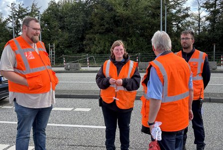 Vier Personen in orangefarbenen Warnwesten stehen auf einer befestigten Fläche am Straßenrand und unterhalten sich. Im Hintergrund befinden sich Bäume, Bauzäune, Poller und Straßenmarkierungen.