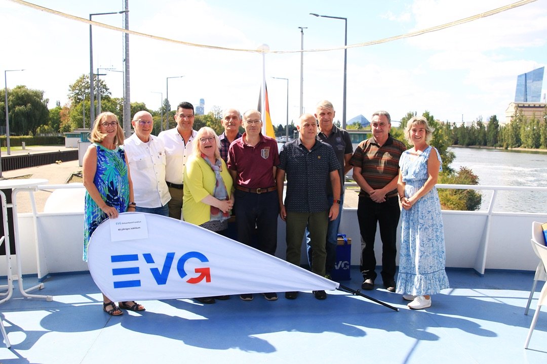 Neun Personen stehen auf dem Außendeck eines Schiffes hinter einer großen EVG-Fahne und posieren für ein Gruppenfoto. Die Gruppe befindet sich auf einem hellblauen Schiffsdeck. Vor den Personen liegt eine weiße Fahne mit dem blauen und roten EVG-Logo. Auf einem kleinen Schild an der Fahne ist der Text „EVG Ortsverband Frankfurt“ sowie „40-jähriges Jubiläum“ zu erkennen. Im Hintergrund steht ein Mast mit einer Deutschlandflagge.