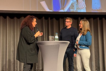 Drei Personen stehen auf einer Bühne vor einem grauen Vorhang an einem runden Stehtisch. Eine Person hält ein Mikrofon. Auf dem Tisch stehen zwei Wasserflaschen, mehrere Gläser und eine Karaffe. Im oberen Bildbereich ist der untere Teil einer Projektionsleinwand sichtbar.