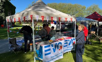 Ein Pavillon im Freien auf einer Rasenfläche mit einem Tisch, der mit einem Banner „EVG – Eisenbahn- und Verkehrsgewerkschaft“ bedeckt ist. Auf dem Tisch liegen Informationsmaterialien und kleine Gegenstände. Am Pavillon hängen rot-weiße Wimpel mit EVG-Logos. Mehrere Personen stehen um den Tisch, im Hintergrund sind weitere Pavillons, Bäume und ein parkähnliches Gelände zu sehen.
