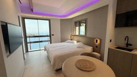 Heller Wohnraum mit Doppelbett, kleinem Esstisch, Wandfernseher und Küchenzeile. Eine große Schiebetür führt auf einen Balkon mit Blick auf das Meer. Indirekte violette Deckenbeleuchtung verläuft entlang einer abgehängten Decke.