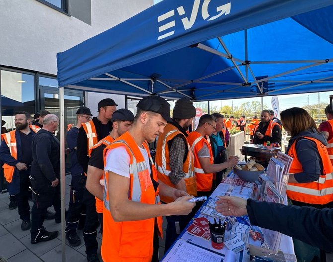 Im Außenbereich vor einem Gebäude steht ein blauer Pavillon mit der weißen Aufschrift „EVG“. Darunter versammeln sich mehrere Personen in orangefarbenen Warnwesten um einen Tisch. Auf dem Tisch liegen gedruckte Informationsmaterialien, Klemmbretter, Stifte und weitere Gegenstände. Im Hintergrund sind weitere Personen, Gebäudefassaden und ein offener Platz zu sehen.