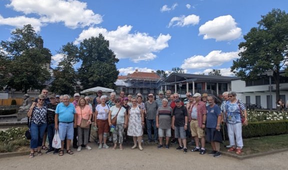 Gruppe von etwa 30 Personen im Freien auf einem hellen Weg vor einem Gebäude mit Säulengang und großen Fenstern. Im Hintergrund stehen Bäume, ein Gebäude mit Gerüst, Sonnenschirme und ein Brunnenbereich. Der Himmel ist blau mit vielen weißen Wolken.