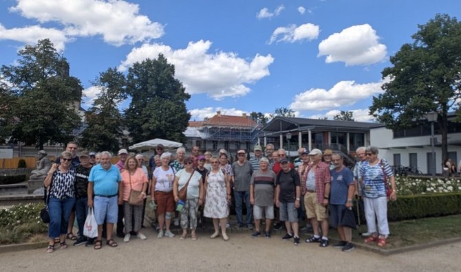 Gruppe von etwa 30 Personen im Freien auf einem hellen Weg vor einem Gebäude mit Säulengang und großen Fenstern. Im Hintergrund stehen Bäume, ein Gebäude mit Gerüst, Sonnenschirme und ein Brunnenbereich. Der Himmel ist blau mit vielen weißen Wolken.