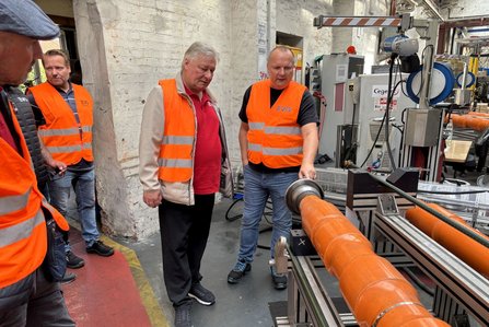 Vier Personen mit orangefarbenen Warnwesten stehen in einer Produktionshalle an einer Maschine. Eine Person zeigt auf ein langes orangefarbenes Rohr, das auf einer Metallvorrichtung liegt. Im Hintergrund sind weitere Maschinenteile, Kabel, Metallrahmen und eine helle Mauer mit sichtbarer Struktur zu sehen.