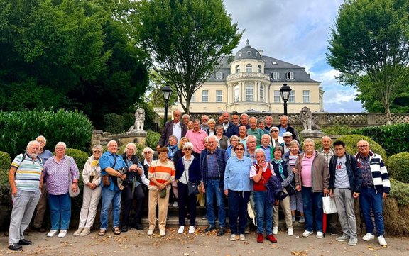 Gruppenfoto von mehreren Personen auf einer Treppe in einem gepflegten Garten. Im Hintergrund steht ein großes, helles Gebäude mit Rundturm und Balkon, eingerahmt von Bäumen und Steinfiguren.