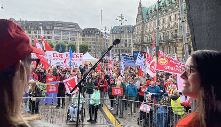 Blick von einer Bühne auf eine große Menschenmenge auf einem gepflasterten Platz vor einem historischen Gebäude. Im Vordergrund stehen ein Mikrofonständer und zwei teilweise sichtbare Personen am Bildrand. In der Menge sind zahlreiche rote, weiße und blau-weiße Fahnen sowie Transparente zu sehen. Auf einem großen Transparent steht gut lesbar „EURE KRIEGE NICHT AUF UNSEREN RÜCKEN!“. Absperrgitter trennen den Bühnenbereich von den Teilnehmenden.