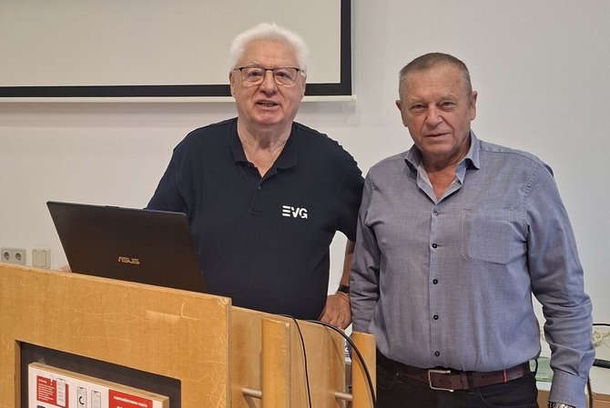 Nahaufnahme des Bereichs vor der Leinwand. Zwei Personen stehen nebeneinander hinter einem hölzernen Rednerpult. Auf dem Pult steht ein aufgeklappter Laptop mit sichtbarer Markenlogo „ASUS“. Auf dem Poloshirt einer Person ist ein „EVG“-Logo zu sehen. Im Hintergrund ist der untere Rand der Projektionsleinwand sowie eine helle Wand mit Steckdosen sichtbar.