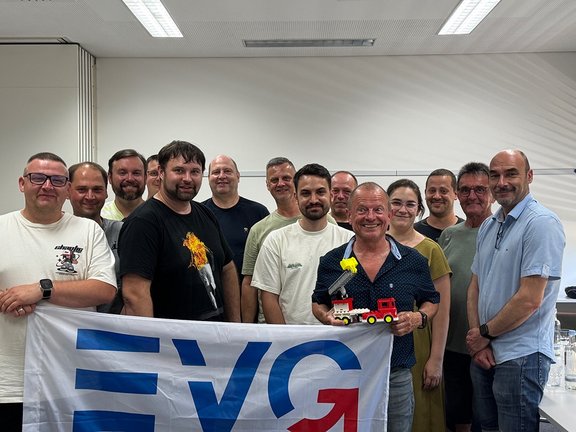 Eine Gruppe von Personen steht in einem Seminarraum um ein EVG‑Banner, während eine Person vorne ein kleines Feuerwehr‑Spielzeugfahrzeug in der Hand hält. Mehrere Personen stehen dicht nebeneinander und schauen in Richtung Kamera. Vorne ist ein großes Banner mit den Buchstaben „EVG“ zu sehen. Eine Person steht im Vordergrund und hält ein kleines rotes Feuerwehrauto mit Leiter hoch. Die übrigen Personen stehen dahinter in einer Reihe und tragen Freizeitkleidung wie T‑Shirts und Hemden.