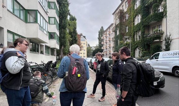Mehrere Personen stehen auf einem Gehweg in einer Wohnstraße zwischen mehrstöckigen Gebäuden. Fahrräder sind am linken Straßenrand abgestellt, rechts stehen geparkte Autos. Der Himmel ist bewölkt. Ein Gebäude auf der rechten Seite ist teilweise mit Kletterpflanzen bewachsen.