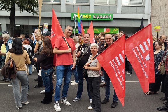 Mehrere Personen stehen bei einer Demonstration auf einer Straße und halten rote Fahnen der Eisenbahn- und Verkehrsgewerkschaft (EVG). Im Vordergrund posiert eine Gruppe von Personen mit mehreren roten Fahnen, auf denen das EVG-Logo sowie der Schriftzug „Eisenbahn- und Verkehrsgewerkschaft“ zu sehen sind. Die Personen befinden sich inmitten einer größeren Menschenmenge. Einige weitere Fahnen und Schilder sind im Hintergrund erkennbar.