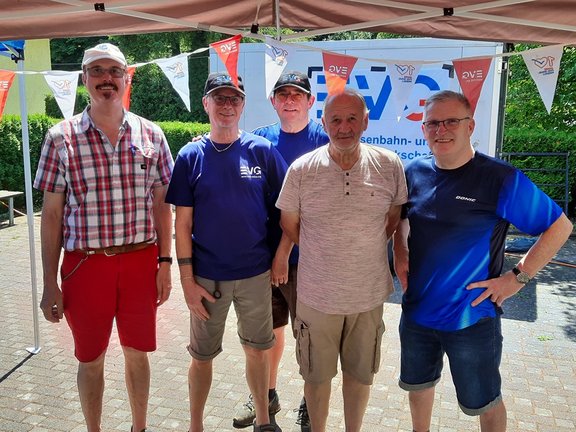 Fünf Personen stehen nebeneinander unter einem Pavillon vor einem EVG‑Banner und blicken in die Kamera. Die fünf Personen stehen dicht beieinander auf einem gepflasterten Bereich im Freien. Über den Köpfen spannt sich ein heller Pavillon mit bunten Wimpeln, auf denen unter anderem „EVG“ zu sehen ist. Hinter der Gruppe hängt ein großes Banner mit der Aufschrift „EVG“ sowie weiteren kleineren Texten. Mehrere Personen tragen Freizeitkleidung wie T‑Shirts, kurze Hosen und Kappen; auf einigen Kleidungsstücken ist ebenfalls „EVG“ zu erkennen. Im Hintergrund sind Bäume und ein Gebäude sichtbar, was auf eine Veranstaltung im Freien hinweist.