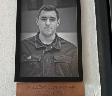 Schwarz-weißes gerahmtes Porträt einer Person in Arbeitskleidung mit DB-Logo an einer hellen Wand. Darunter befindet sich eine Gedenktafel mit der Aufschrift: „In stillem Gedenken an Danny Preuß“.