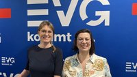 Zwei Personen stehen vor einer blauen Fotowand mit mehrfach sichtbarem weißen Schriftzug „EVG kommt an.“. Die linke Person trägt ein dunkles Oberteil und eine Brille. Die rechte Person trägt eine helle Bluse mit floralem Muster und eine Brille. Beide sind von der Hüfte aufwärts zu sehen.