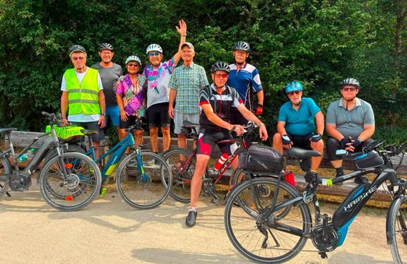 Neun Personen mit Fahrradhelmen posieren mit mehreren Fahrrädern auf einem befestigten Weg vor dichtem grünen Baumbestand. Einige Personen stehen, zwei sitzen auf einer Holzbank. Im Vordergrund stehen und lehnen mehrere Fahrräder, darunter ein schwarzes E‑Bike rechts und weitere Fahrräder links und in der Mitte. Eine Person in der Bildmitte hebt einen Arm nach oben. Die Szene ist bei Tageslicht aufgenommen.