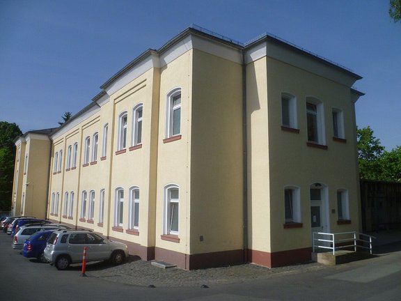 Zweigeschossiges, hellgelbes Gebäude mit weißen Fensterrahmen und rotbraunen Fensterbänken. Entlang der linken Gebäudeseite stehen mehrere geparkte Autos auf einem gepflasterten und asphaltierten Parkplatz. An der rechten Gebäudeecke befindet sich ein Eingang mit einer kleinen Rampe und weißem Geländer. Das Gebäude wird von Tageslicht beleuchtet; im Hintergrund sind Bäume und ein blauer Himmel zu sehen.
