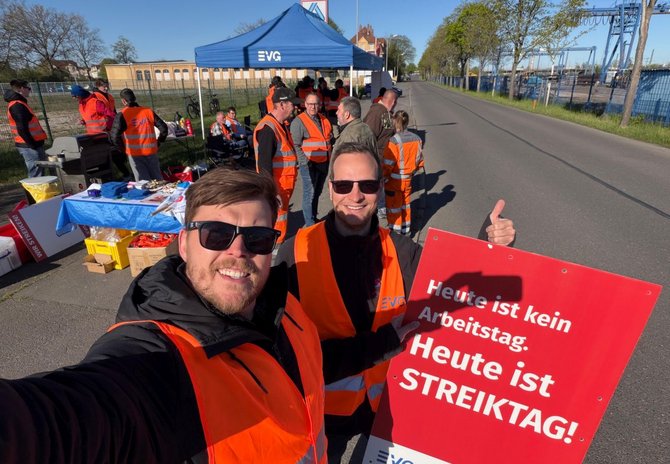 Auf einer Straße stehen mehrere Menschen in orange Warnwesten. Im Vordergrund hält eine Person ein rotes Schild mit der Aufschrift „Heute ist kein Arbeitstag. Heute ist STREIKTAG!“. Im Hintergrund sind ein blauer Pavillon mit EVG‑Logo, Tische mit Gegenständen und weitere Personen zu sehen.