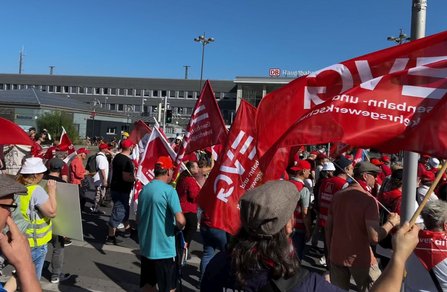 Seitliche Ansicht einer Demonstration mit zahlreichen Personen, viele tragen rote Kleidung oder halten rote Fahnen mit „EVG“-Aufdruck. Im Hintergrund ist erneut das Gebäude mit der Beschriftung „DB Hauptbahnhof“ sichtbar. Die Menge bewegt sich entlang einer Straße. Straßenlaternen, Verkehrszeichen und ein klarer Himmel sind erkennbar.