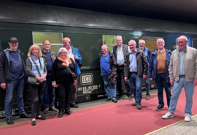 Gruppe von etwa zwölf Personen steht in einer Reihe auf einem Bahnsteig vor einem dunkelgrünen Eisenbahnwagen. Die Personen tragen Jacken, Pullover, Hemden und Jeans. Der Boden des Bahnsteigs ist rot und grau. Am Wagen sind weiße Beschriftungen und Logos sichtbar, darunter „DB“ sowie Zahlen und Buchstaben wie „80 89-80 305-1“ und „WGSüge“. Im Hintergrund ist die Seitenwand des Wagens mit Fenstern zu sehen, die Umgebung wirkt wie ein überdachter oder unterirdischer Bahnhof.