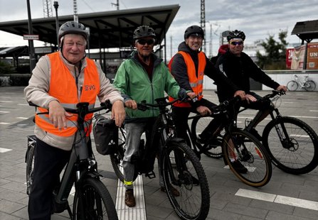 Vier Personen stehen mit Fahrrädern auf einem gepflasterten Platz im Außenbereich eines Bahnhofs. Zwei Personen tragen orangefarbene Warnwesten mit sichtbarem „EVG“-Schriftzug, alle tragen Fahrradhelme. Im Hintergrund sind Bahnsteigdächer, Oberleitungsmasten, Schienenanlagen und mehrere abgestellte Fahrräder zu sehen. Der Himmel ist bewölkt.